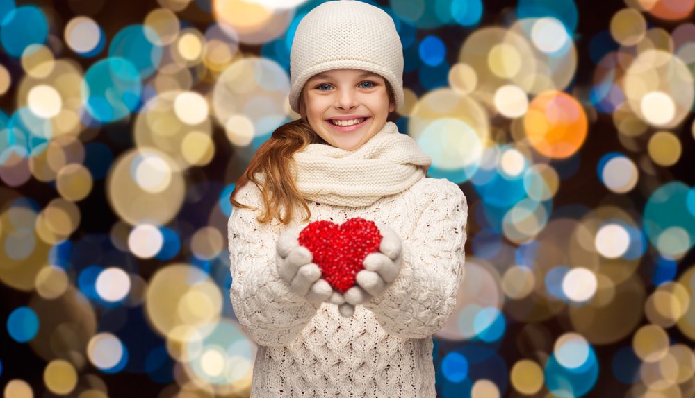 Une petite fille souriante tient un cœur rouge dans ses mains devant des lumières floues.