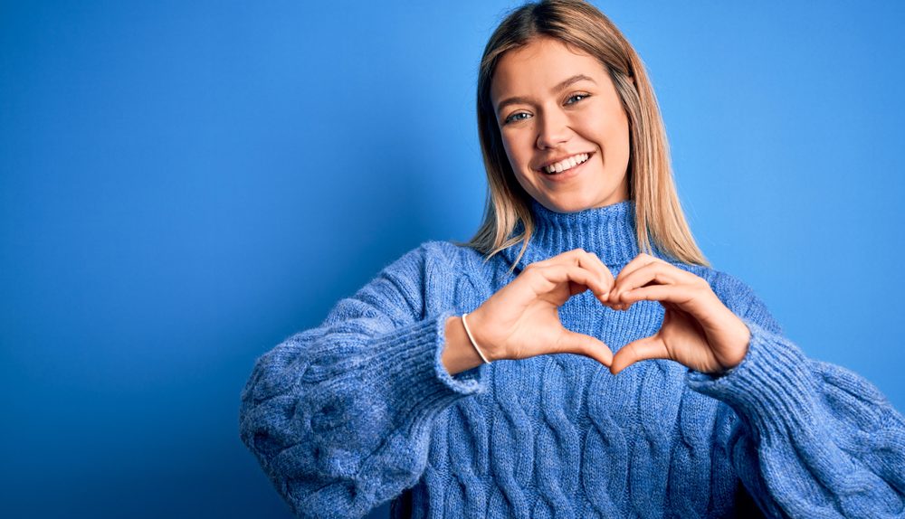 Une jeune femme souriante forme un cœur avec ses mains devant un fond bleu.