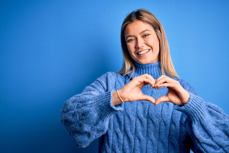 Une jeune femme souriante forme un cœur avec ses mains devant un fond bleu.