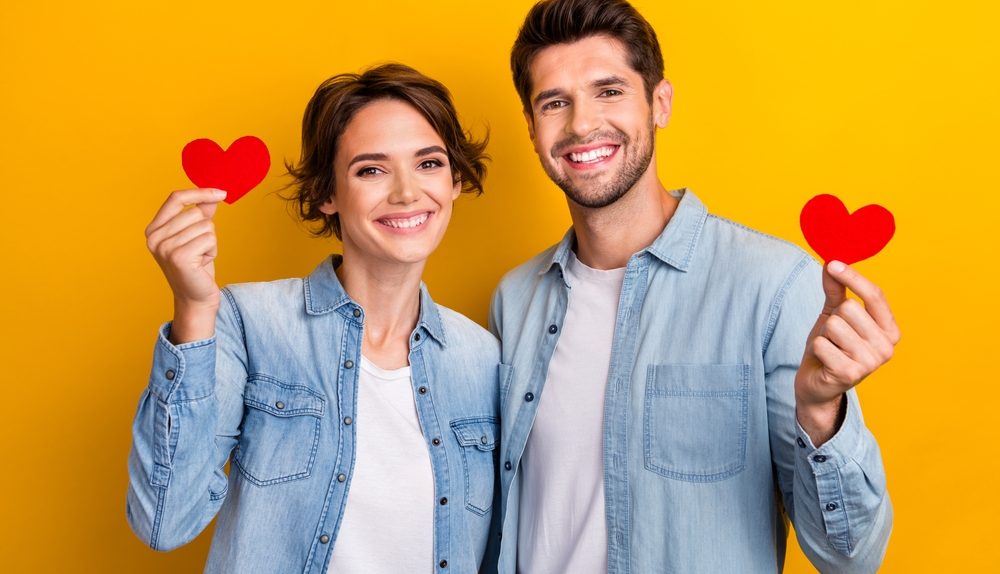 Un homme et une femme souriants tiennent chacun un petit cœur rouge devant un fond jaune.