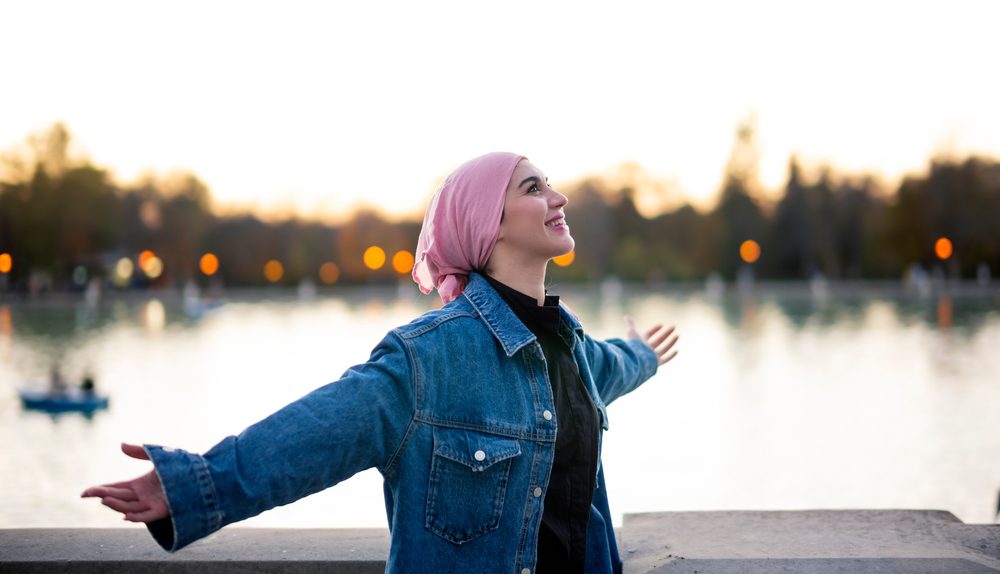 Jeune femme souriante les bras ouverts au bord d’un lac au coucher du soleil.