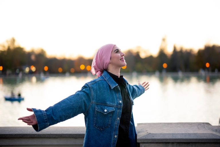 Jeune femme souriante les bras ouverts au bord d’un lac au coucher du soleil.