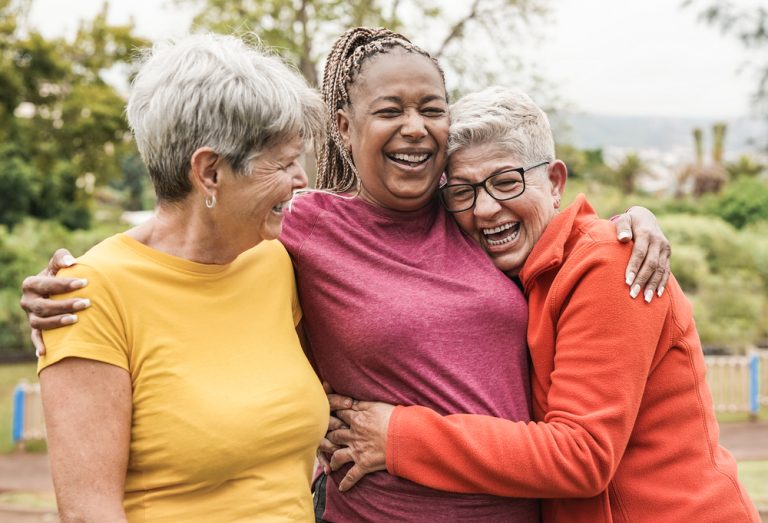 Trois femmes souriantes se prennent dans les bras en extérieur.