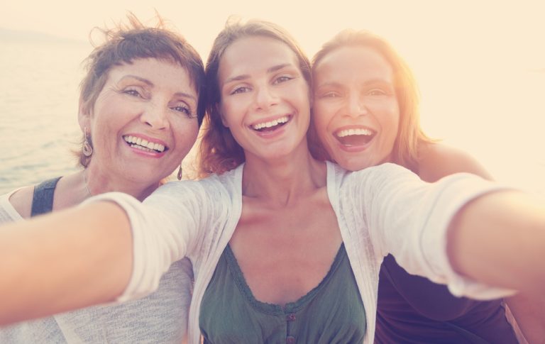 Trois femmes souriantes prenant un selfie ensemble au coucher du soleil.
