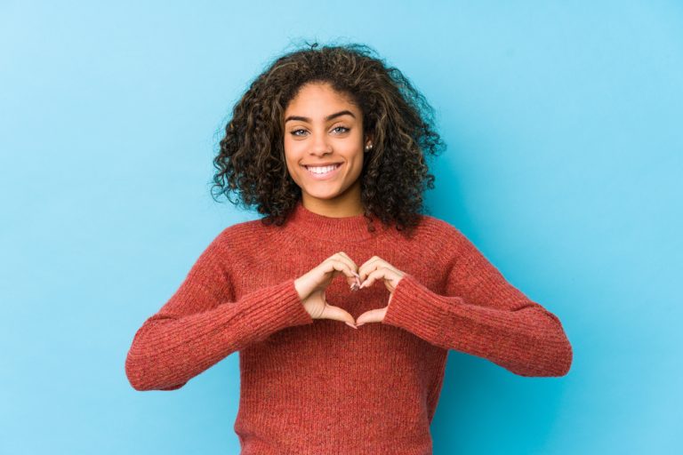 Jeune femme souriante formant un cœur avec ses mains sur un fond bleu.