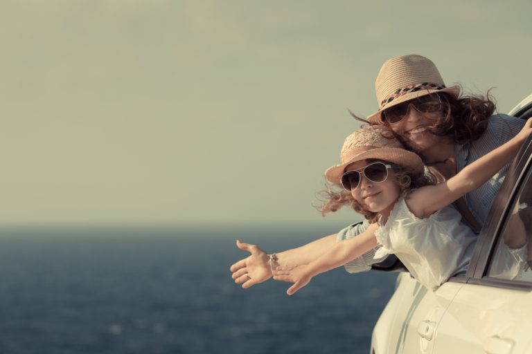 Mère et enfant souriants sortant d’une voiture près de la mer pour symboliser les vacances, le bien-être familial et la liberté.