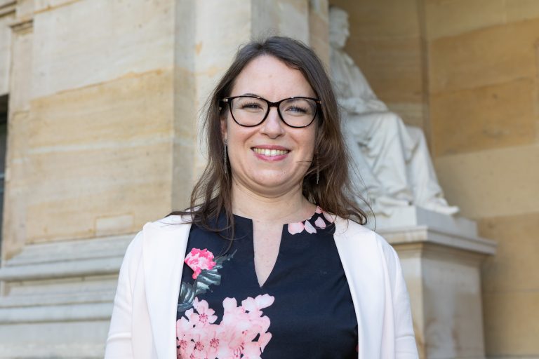 Portrait d’une femme souriante portant des lunettes lors d’un événement officiel dans un cadre institutionnel.