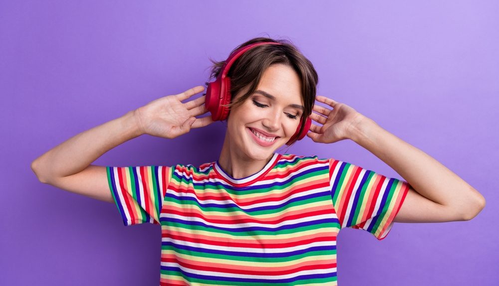 Jeune femme souriante écoutant de la musique avec un casque audio sur un fond violet.