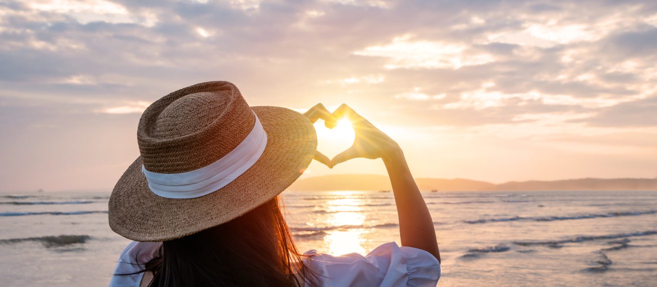 Femme face à la mer formant un cœur avec ses mains au coucher du soleil pour symboliser le bien-être, l’espoir et la santé du cœur.