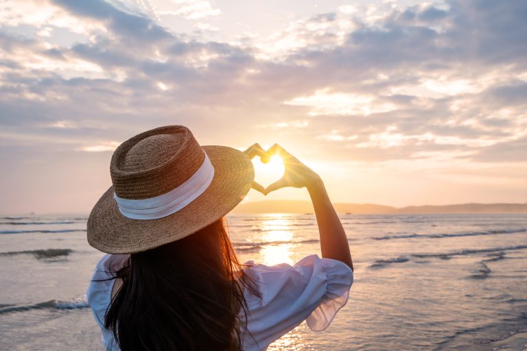 Femme face à la mer formant un cœur avec ses mains au coucher du soleil pour symboliser le bien-être, l’espoir et la santé du cœur.