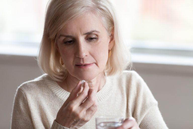 Femme mature tenant un comprimé et un verre d’eau pour illustrer la prise de traitement médical et le suivi de la santé.