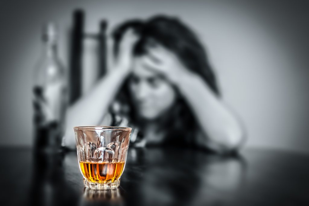 Verre d’alcool au premier plan avec une femme en détresse en arrière-plan pour illustrer les dangers de l’alcool et des addictions.