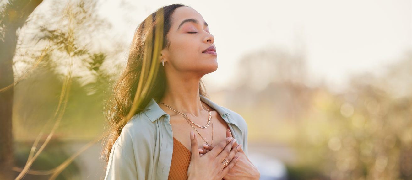Jeune femme les yeux fermés tenant ses mains sur sa poitrine dans un moment de calme et de bien-être en pleine nature.