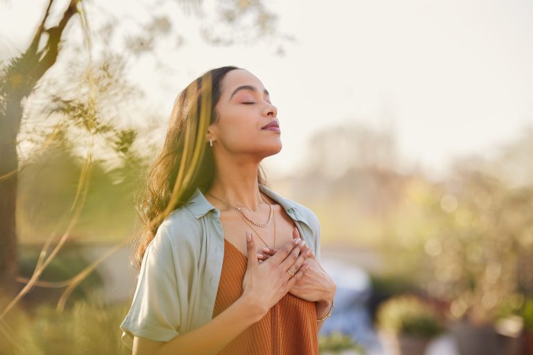 Jeune femme les yeux fermés tenant ses mains sur sa poitrine dans un moment de calme et de bien-être en pleine nature.