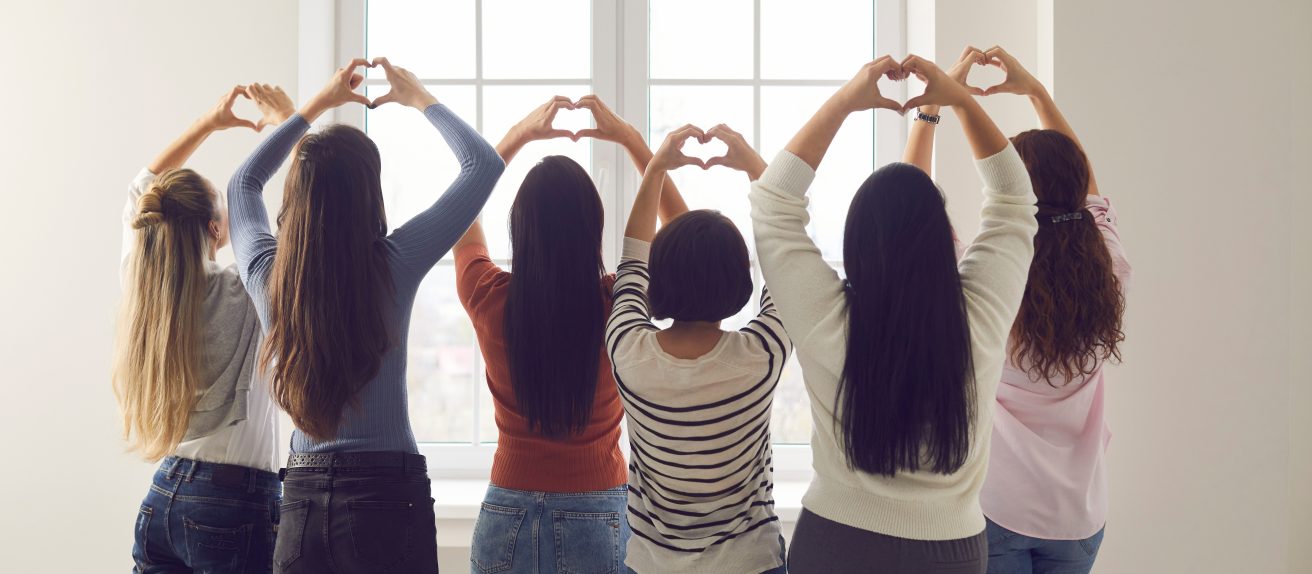 Groupe de femmes formant des cœurs avec leurs mains devant une fenêtre pour symboliser la solidarité, le soutien et la sensibilisation à la santé des femmes.