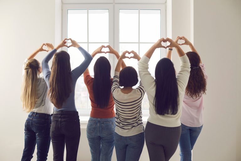 Groupe de femmes formant des cœurs avec leurs mains devant une fenêtre pour symboliser la solidarité, le soutien et la sensibilisation à la santé des femmes.