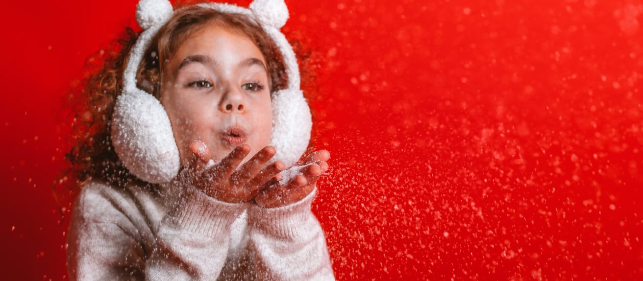 Petite fille soufflant des flocons de neige sur fond rouge de Noël pour symboliser la joie, l’espoir et la santé du cœur pendant les fêtes