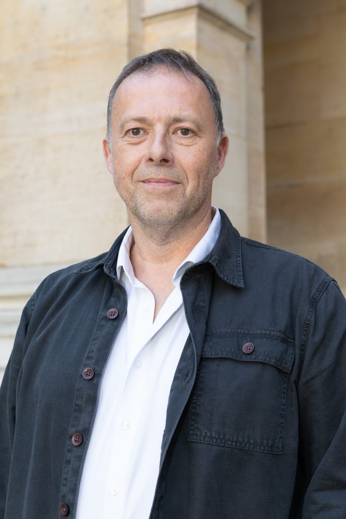 Portrait d’un homme portant une veste sombre et une chemise blanche devant un bâtiment en pierre claire.