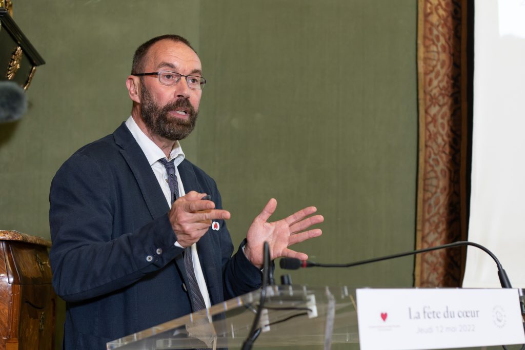 Portrait rapproché d’un homme barbu avec des lunettes, vêtu d’un costume bleu et d’une cravate grise, en train de parler.