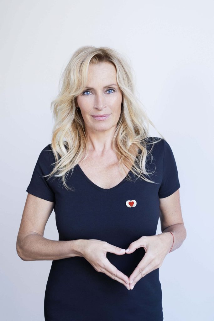 Portrait d’une femme blonde portant un t-shirt noir avec un symbole de cœur et formant un cœur avec ses mains.