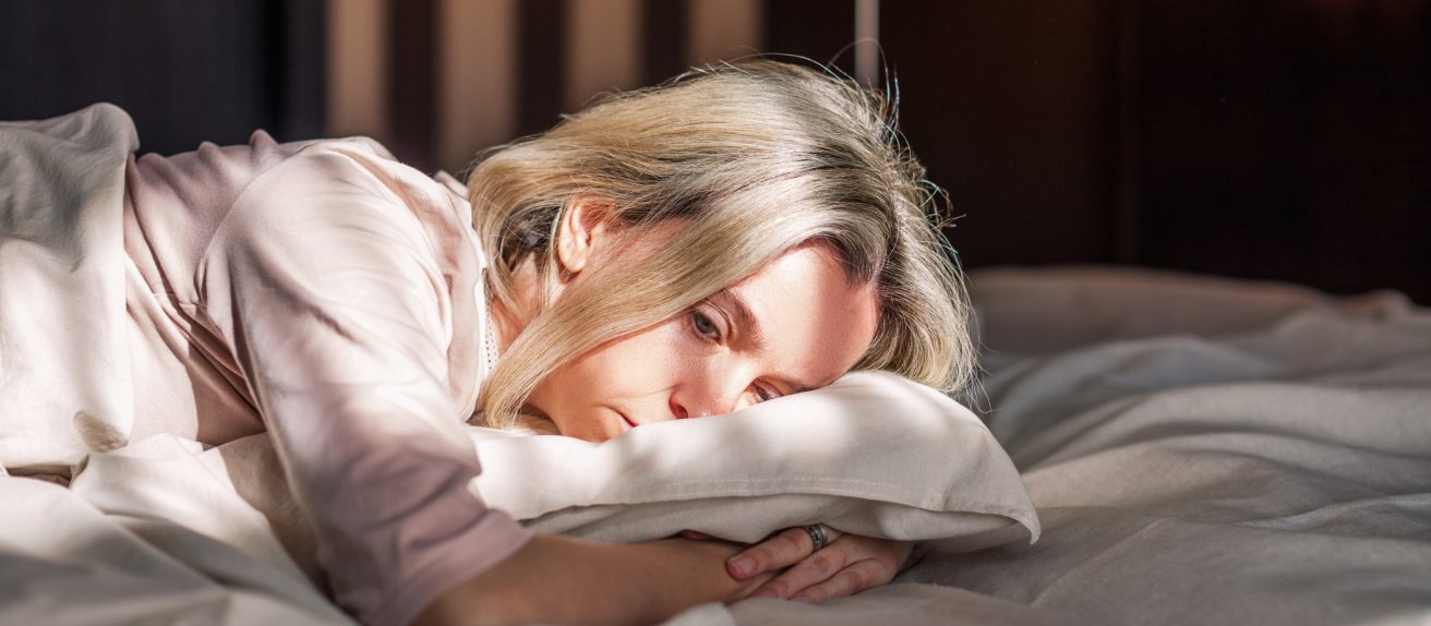 Une femme blonde allongée sur un lit regarde dans le vide avec un air fatigué et pensif.