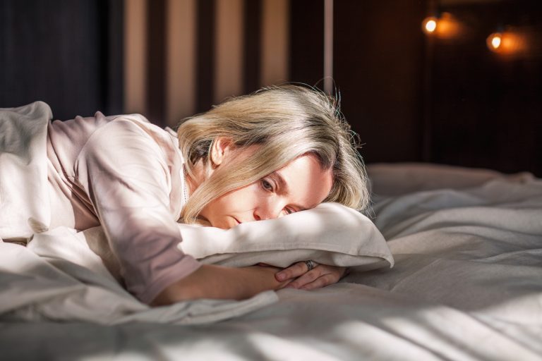 Une femme blonde allongée sur un lit regarde dans le vide avec un air fatigué et pensif.