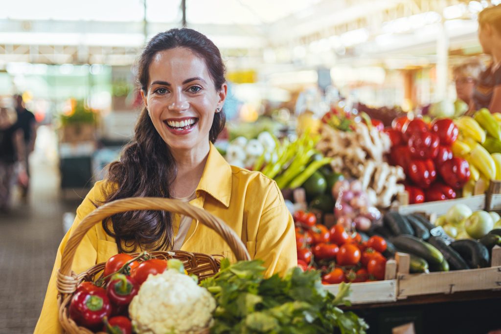 Une femme souriante tient un panier de légumes dans un marché.