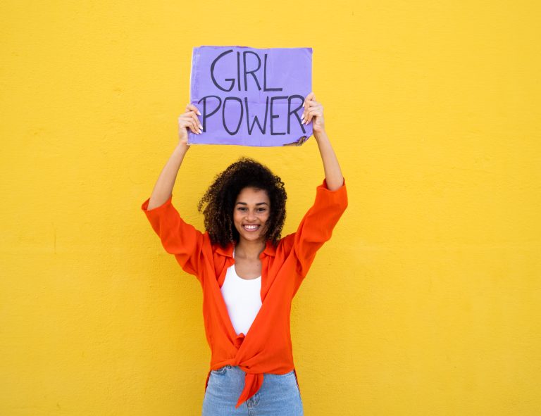 Jeune femme tenant une pancarte “Girl Power” devant un mur jaune.